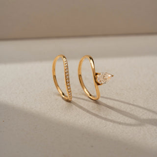 Angled view of a pear solitaire ring in 14 KT Yellow Gold, featuring a bezel-set pear-shaped lab-grown diamond paired with a pavé-set curved band in a modern stacked design, displayed against a beige backdrop.
