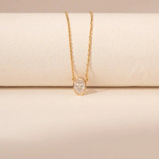 Close-up view of a 14KT yellow gold necklace featuring an oval-cut solitaire diamond pendant, set against a beige background.