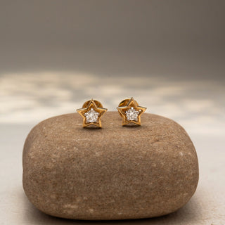 Front view of star-shaped diamond stud earrings in 14 KT Yellow Gold, featuring open star frames with brilliant round center diamonds, displayed on a textured stone surface.
