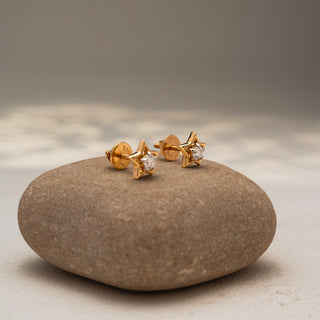 Side view of star-shaped diamond stud earrings in 14 KT Yellow Gold, featuring open star frames with brilliant round center diamonds, displayed on a textured stone surface.