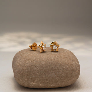 Angled view of star-shaped diamond stud earrings in 14 KT Yellow Gold, featuring open star frames with brilliant round center diamonds, displayed on a textured stone surface.
