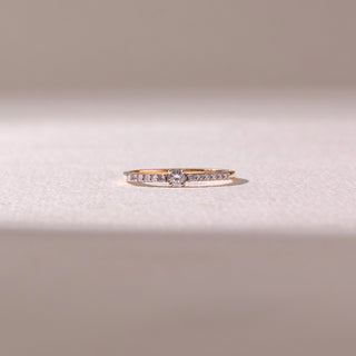 Front view of a delicate yellow gold diamond ring, featuring a round center stone with pavé-set band detailing. Captured against a soft beige backdrop, the design reflects minimal elegance and sparkle.
