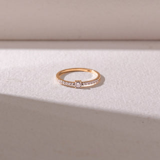 Top view of a delicate yellow gold diamond ring, featuring a round center stone with pavé-set band detailing. Captured against a soft beige backdrop, the design reflects minimal elegance and sparkle.