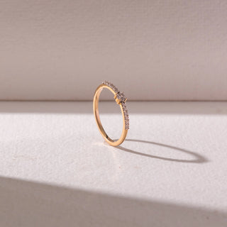 Angled view of a delicate yellow gold diamond ring, featuring a round center stone with pavé-set band detailing. Captured against a soft beige backdrop, the design reflects minimal elegance and sparkle.