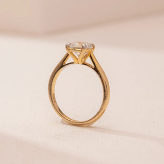 Side view of Reia’s 14 KT Rose Gold Golden Promise Diamond Solitaire Ring. The round diamond is held in a tall, open setting on a smooth band, shown on a soft pink surface.
