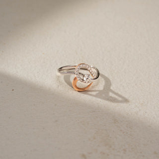 Top view of a two-tone infinity diamond ring in 14 KT Gold, featuring a round lab-grown diamond nestled within intertwined rose and white gold loops accented with pavé-set diamonds, displayed against a beige backdrop