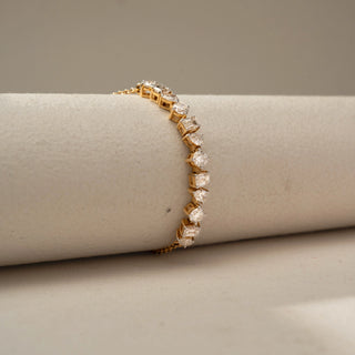 Side view of a gold bracelet featuring alternating round, pear, and emerald-cut gemstones, highlighting the profile of each setting on a neutral cylindrical display stand.