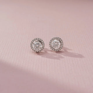 Front view of a pair of round halo stud earrings with sparkling center stones surrounded by pavé accents, displayed on a soft pink surface with gentle natural light and shadows.