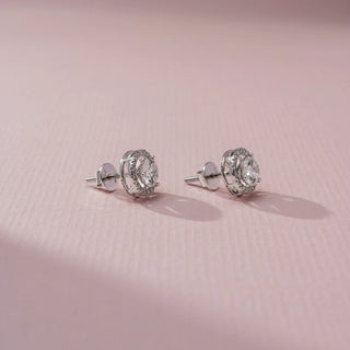 Side view of a pair of halo stud earrings featuring round center stones surrounded by pavé accents, with screw-back posts, displayed on a soft pink surface in gentle light.