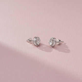 Angled side view of a pair of halo diamond stud earrings with round center stones and pavé-set halos, featuring screw-back posts, displayed on a soft pink textured surface.