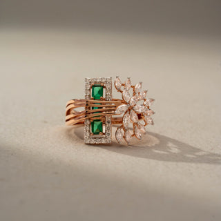 Front view of a floral statement ring in 14 KT Rose Gold, featuring emerald-cut green gemstones framed by diamond halos, accented with marquise-cut lab-grown diamonds in a blooming floral motif, displayed against a beige backdrop.