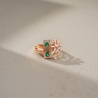 Side view of a floral statement ring in 14 KT Rose Gold, featuring emerald-cut green gemstones framed by diamond halos, accented with marquise-cut lab-grown diamonds in a blooming floral motif, displayed against a beige backdrop.