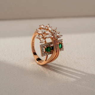 Angled view of a floral statement ring in 14 KT Rose Gold, featuring emerald-cut green gemstones framed by diamond halos, accented with marquise-cut lab-grown diamonds in a blooming floral motif, displayed against a beige backdrop.
