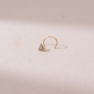 Angled view of a yellow gold nose pin, featuring a floral cluster of round diamonds in a minimal setting. Captured against a soft beige backdrop, the design reflects delicate sparkle and elegance.

