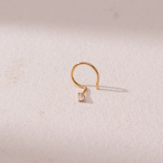 Top view of a yellow gold nose pin, featuring a solitaire round diamond in a minimal prong setting. Captured against a soft beige backdrop, the design reflects delicate sparkle and understated elegance.