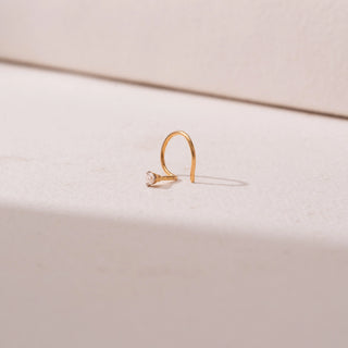 Side view of a yellow gold nose pin, featuring a solitaire round diamond in a minimal prong setting. Captured against a soft beige backdrop, the design reflects delicate sparkle and understated elegance.