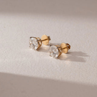 Side view of diamond stud earrings in 14KT yellow gold, featuring round brilliant diamonds in prong settings with screw backs, displayed on a beige surface highlighting classic brilliance and detail.

