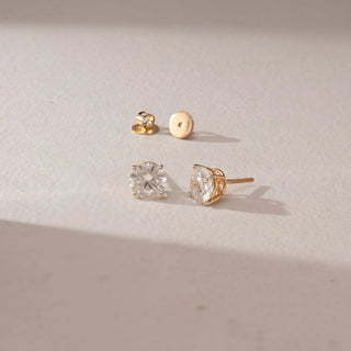 Angled view of diamond stud earrings in 14KT yellow gold, featuring round brilliant diamonds in prong settings with screw backs, placed on a beige surface highlighting detail and sparkle.