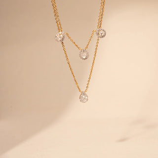 Close up view of a pear-shaped lab-grown diamond layered necklace in 14KT Yellow Gold, featuring halo-set pear and round motifs. Captured against a beige backdrop, the layered design exudes refined elegance.