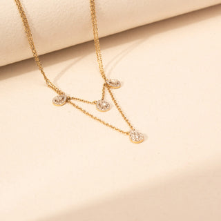 Angled view of a round lab-grown diamond layered necklace in 14KT Yellow Gold, featuring halo-set round motifs with cascading chains. Captured against a beige backdrop, the layered design offers graceful sparkle.