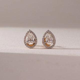 Front view of diamond stud earrings in 14KT yellow gold, featuring pear-shaped center diamonds surrounded by pavé halo settings, displayed on a beige surface highlighting brilliance and elegant design.
