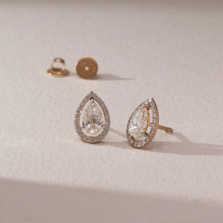 Angled view of diamond stud earrings in 14KT yellow gold, featuring pear-shaped center diamonds with pavé halo settings and screw backs, placed on a beige surface highlighting brilliance and fine detail.
