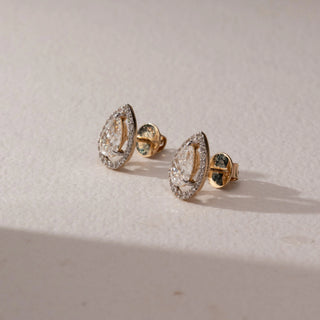 Side view of diamond stud earrings in 14KT yellow gold, featuring pear-shaped center diamonds with pavé halo settings and screw backs, set on a beige surface highlighting depth and brilliance.
