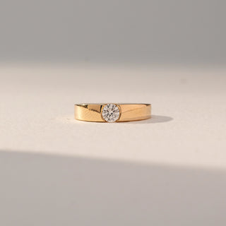 Front view of Bezel Set Diamond Band Ring in 14KT Yellow Gold, featuring a single round lab grown diamond set in a smooth band. Set on a beige backdrop, the design highlights minimal elegance and clean finish.
