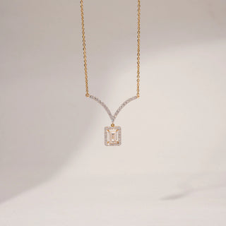 Front view of an emerald-cut lab-grown diamond pendant necklace in 14KT Dual Gold, featuring a halo-set emerald-cut centre stone suspended from a curved diamond bar. Captured against a beige backdrop, the elegant design radiates brilliance.