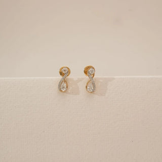Front view of infinity loop diamond earrings featuring round and pear-shaped lab-grown diamonds set in a flowing gold curve, displayed on a neutral beige surface with soft studio lighting.