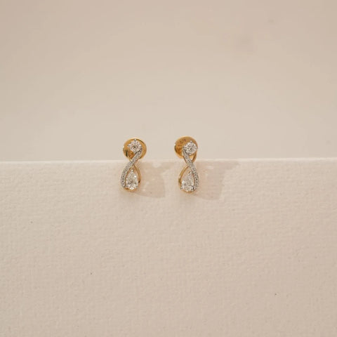 Front view of infinity loop diamond earrings featuring round and pear-shaped lab-grown diamonds set in a flowing gold curve, displayed on a neutral beige surface with soft studio lighting.