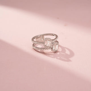 Angled view of a double halo wrap ring in White Gold, featuring two emerald-cut lab-grown diamonds framed by sparkling halo settings on an open-ended pavé-set band, displayed against a soft pink backdrop.