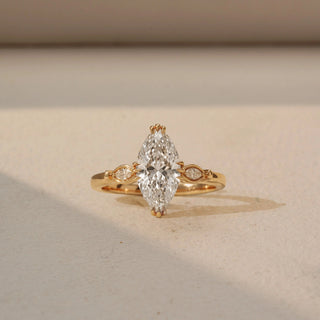 Front view of a marquise solitaire engagement ring in 14 KT Yellow Gold, featuring a marquise-cut lab-grown diamond accented by bezel-set round diamonds within marquise-shaped side motifs, displayed against a beige backdrop.