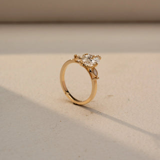 Angled view of a marquise solitaire engagement ring in 14 KT Yellow Gold, featuring a marquise-cut lab-grown diamond accented by bezel-set round diamonds within marquise-shaped side motifs, displayed against a beige backdrop.