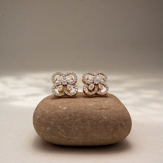 Front view of floral diamond stud earrings in 14 KT Yellow Gold, featuring four petal-inspired pavé diamond loops with pear-shaped diamond accents, displayed on a textured stone surface.
