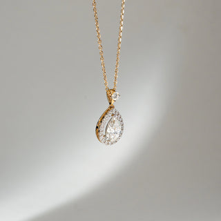 Angled view of Teardrop Halo Diamond Pendant Necklace in 14KT Yellow Gold, featuring a pear-shaped lab grown diamond with halo detailing and a round accent above. Set on a neutral backdrop, the design highlights depth and brilliance.
