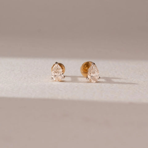 Front view of diamond stud earrings in 14KT yellow gold, featuring pear-shaped diamonds in prong settings, displayed on a beige surface highlighting delicate brilliance and timeless elegance.
