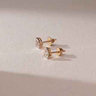 Side view of diamond stud earrings in 14KT yellow gold, featuring pear-shaped diamonds in prong settings with screw backs, displayed on a beige surface highlighting refined brilliance and detail.
