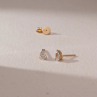 Angled view of diamond stud earrings in 14KT yellow gold, featuring pear-shaped diamonds in prong settings with screw backs, placed on a beige surface highlighting subtle sparkle and fine detail.
