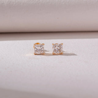 Front view of square diamond stud earrings in yellow gold, featuring solitaire princess-cut diamonds in four-prong settings. Captured against a soft beige backdrop, the design reflects timeless sparkle and elegance.
