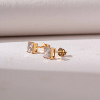 Angled view of square diamond stud earrings in yellow gold, featuring solitaire princess-cut diamonds in four-prong settings. Captured against a soft beige backdrop, the design reflects timeless sparkle and elegance.
