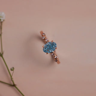 Top view of Reia Blush Accent Azure Diamond Ring on a soft blush-peach background, featuring an oval blue lab-grown diamond and six blush accent stones set in 14KT rose gold.