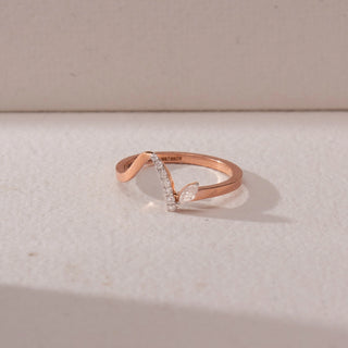 Top view of a diamond ring in 9KT rose gold, featuring a curved pavé band with a marquise diamond accent, set on a beige surface highlighting fluid design and soft sparkle.
