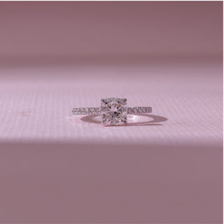 Front view of a cushion-cut diamond engagement ring in white gold, featuring a centred cushion-cut diamond with pavé-set bands. Captured on a soft pink backdrop, highlighting radiant elegance.
