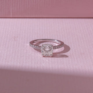Top view of a cushion-cut diamond engagement ring in white gold, featuring a centred cushion-cut diamond with pavé-set bands. Captured on a soft pink backdrop, highlighting timeless sparkle.
