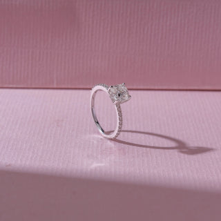 Angled view of a cushion-cut diamond engagement ring in white gold, featuring a centred cushion-cut diamond with pavé-set bands. Captured on a soft pink backdrop, highlighting elegant brilliance.
