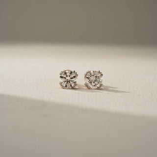 Front view of round brilliant-cut diamond stud earrings in a classic prong setting, displayed on a soft neutral surface with warm natural light and subtle shadows.