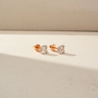 Side view of round brilliant-cut diamond stud earrings in a rose gold prong setting, showcasing the profile, posts, and secure backs on a softly lit neutral surface.