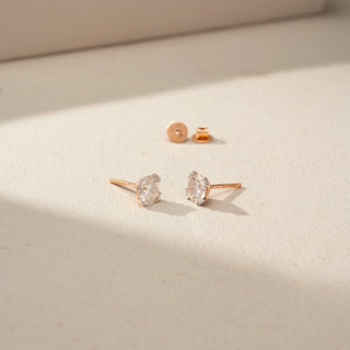 Angled view of round brilliant-cut diamond stud earrings in a rose gold prong setting, showcasing the diamond profile, posts, and detached screw backs on a softly lit neutral surface.