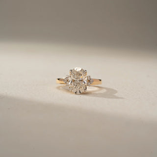 Front view of an oval three-stone engagement ring in 14 KT Yellow Gold, featuring an oval-cut lab-grown diamond flanked by round diamond clusters, displayed against a beige backdrop.